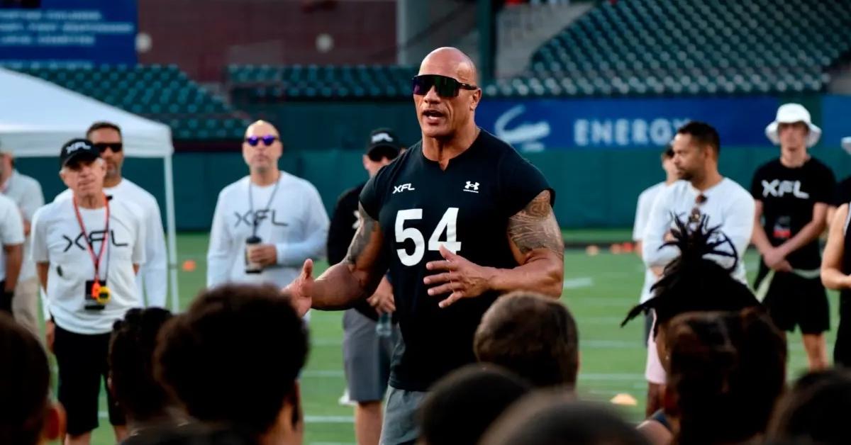 Dwayne Johnson wearing a black XFL shirt speaking with members.