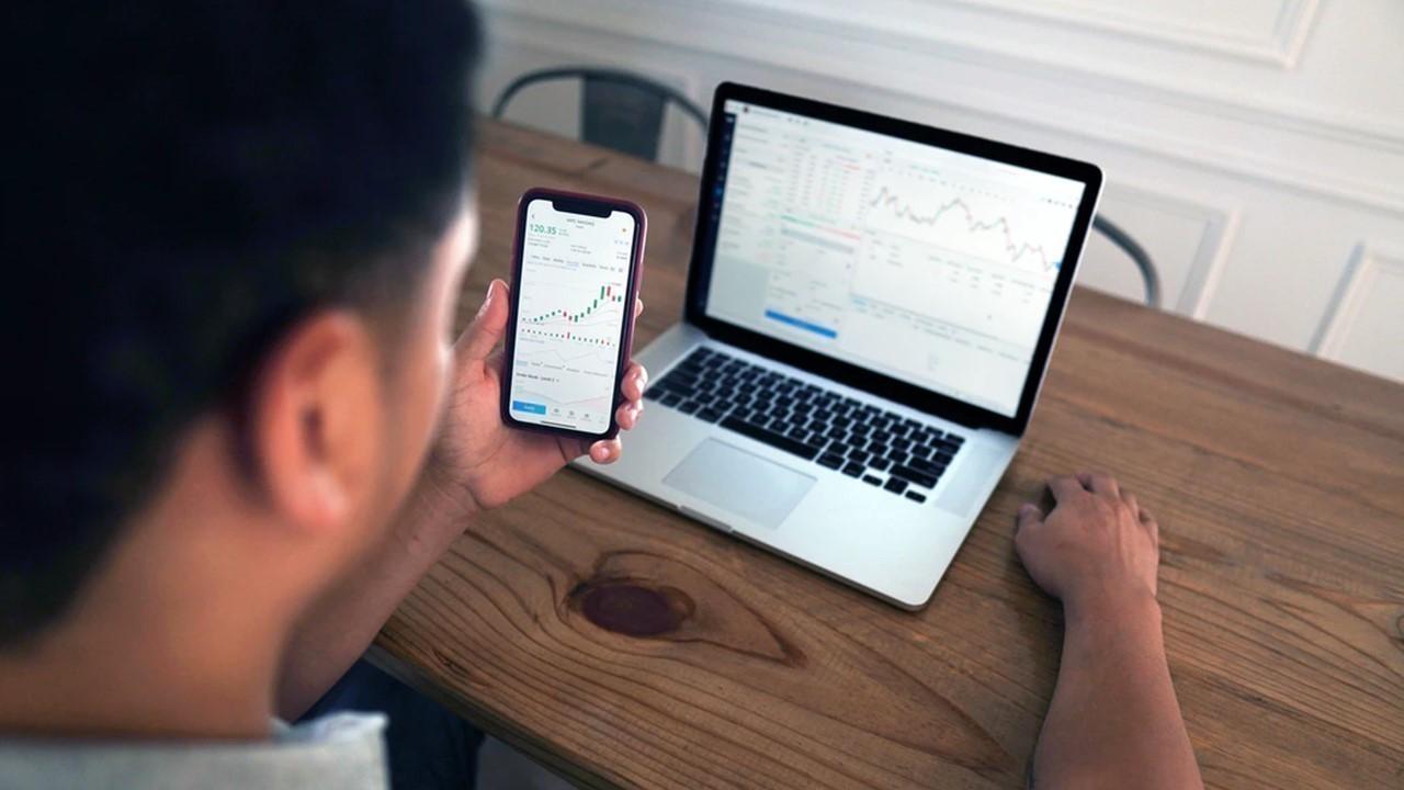 A man looking at stock data on a laptop and smartphone