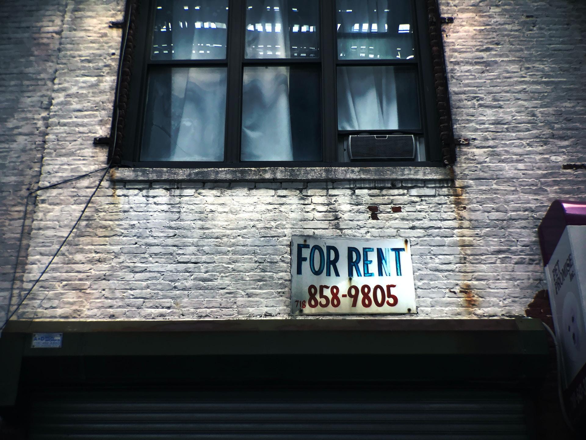 An older apartment building with a "for rent" sign seen on worn white brick