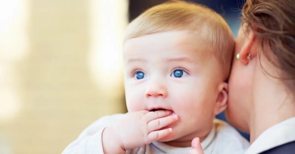 A baby with blue eyes sucks his and while being held by his mom.