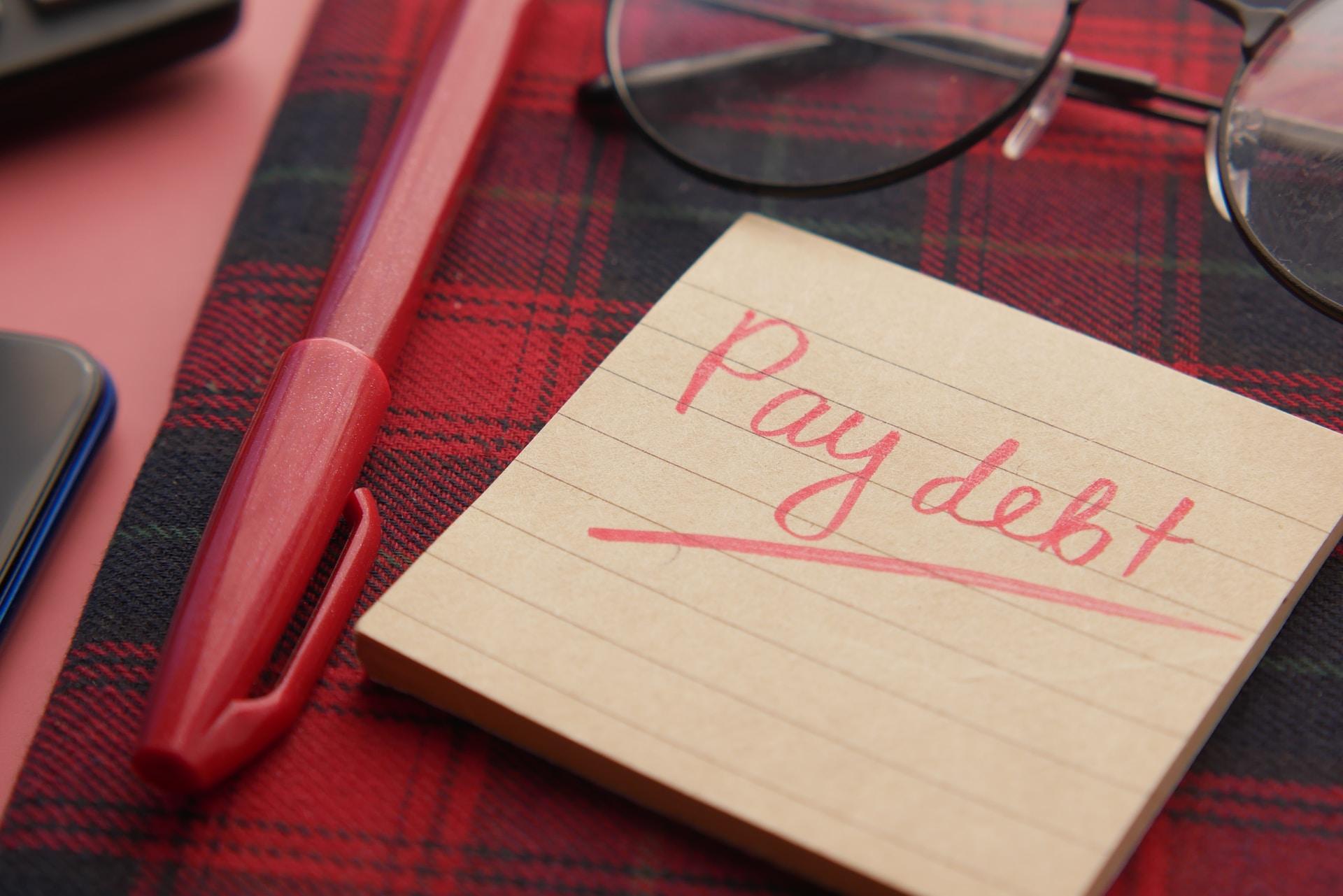 A note pad is seen with the words "pay debt" written across it in red while being underlined