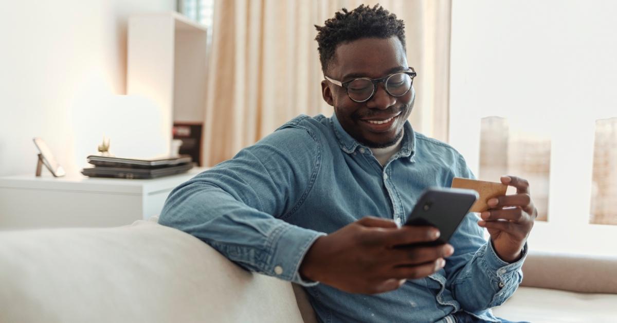 A young man in denim holding his credit card while using a smartphone at home. 