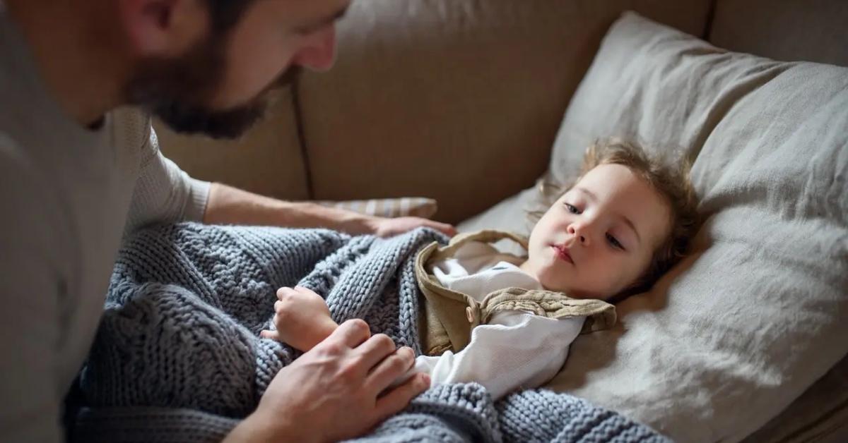 A young sick child is comforted by her dad while she lays on the couch.