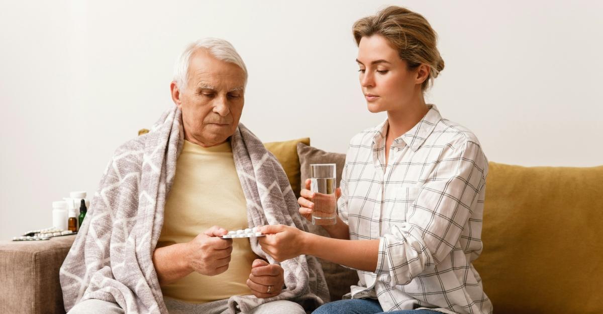 A young woman gives an older man medicine as he sits on the couch with a blanket over his shoulders.
