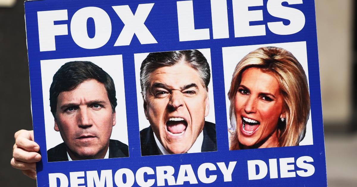 A person holding a sign that says," Fox Lies Democracy Dies" with Fox News anchors on it.