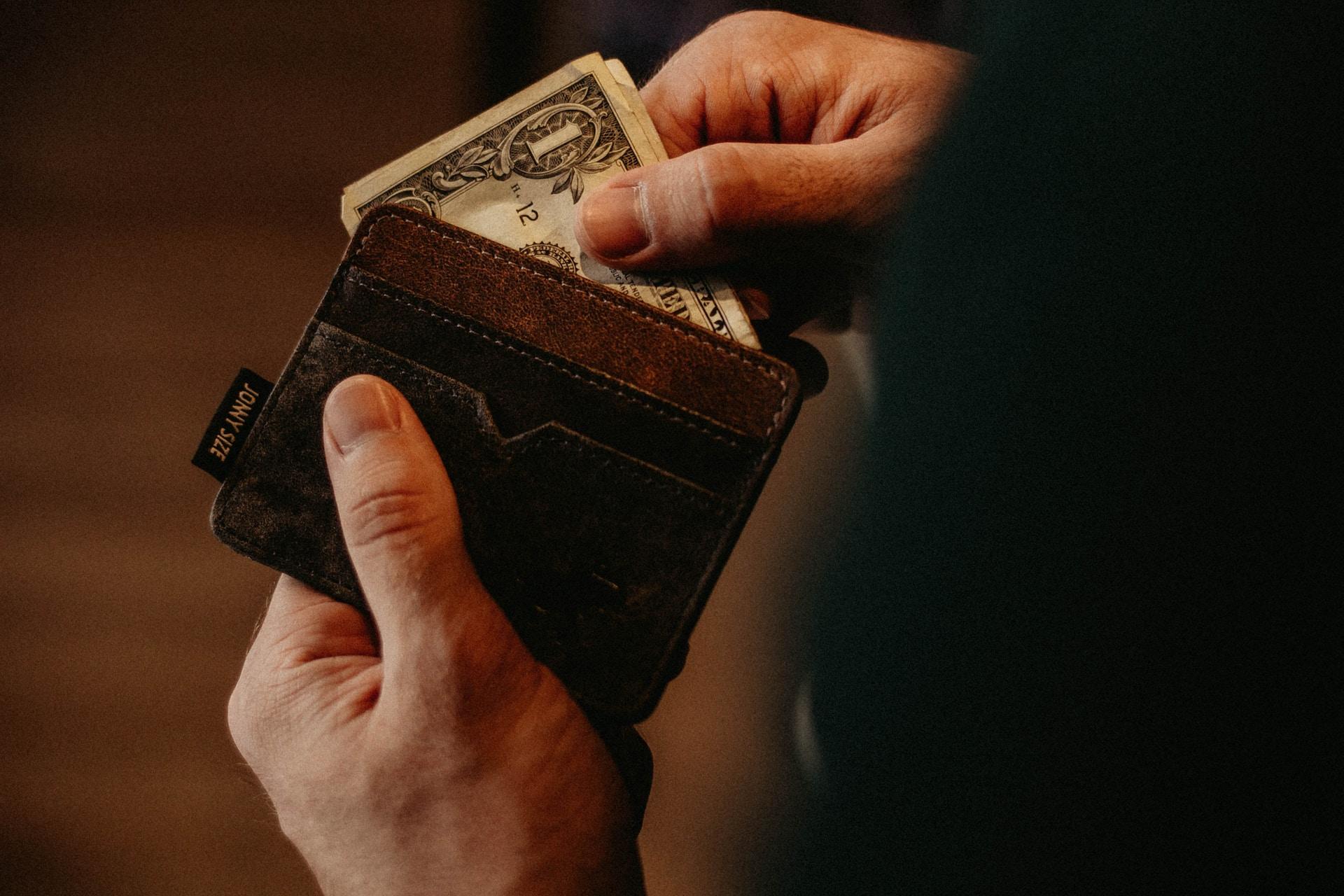 A person holding a brown leather wallet getting a one dollar bill out of it 