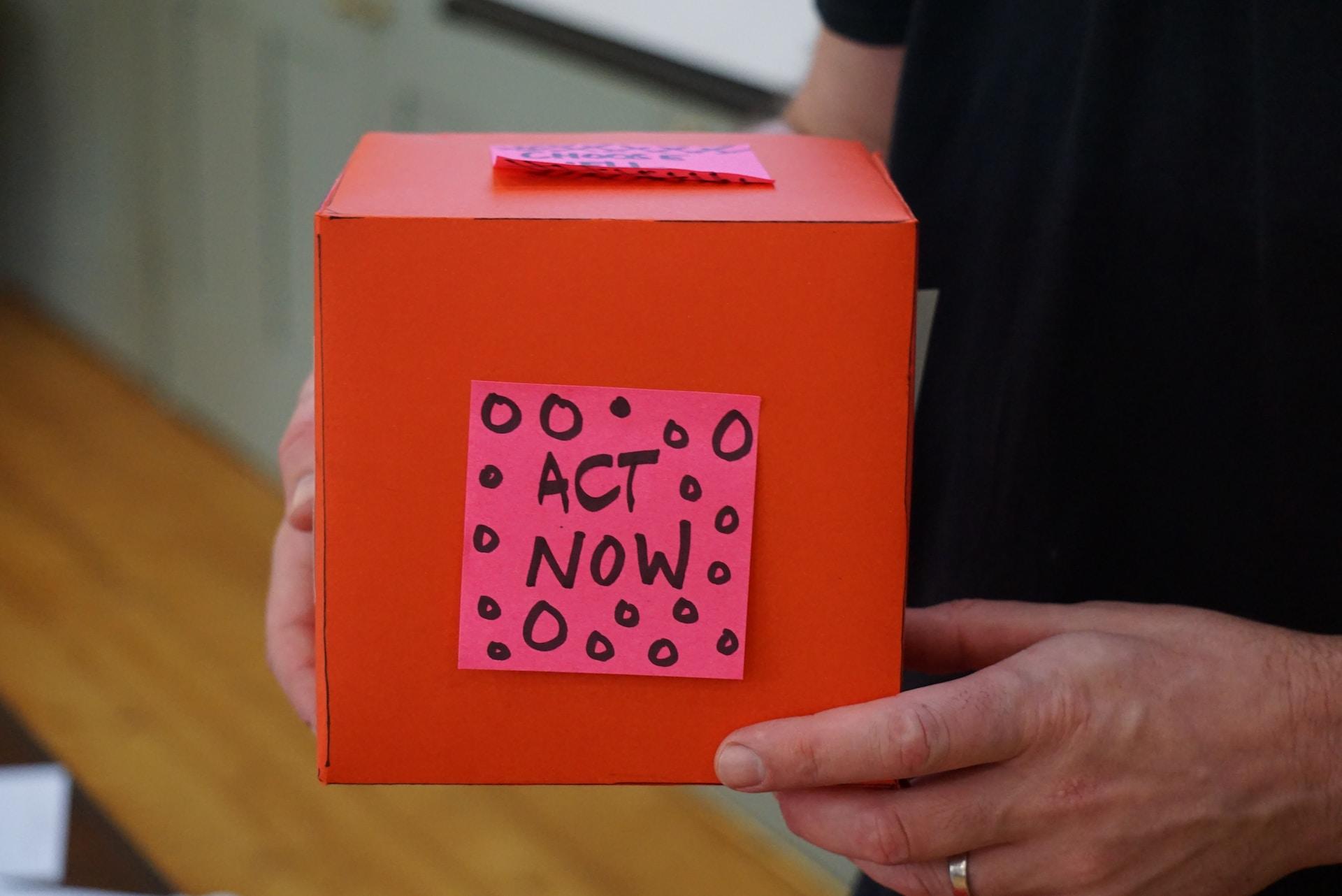 A red donation box with an act now post-it on the side 