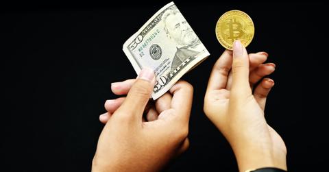 One hand holding a paper bill and another holding a gold Bitcoin