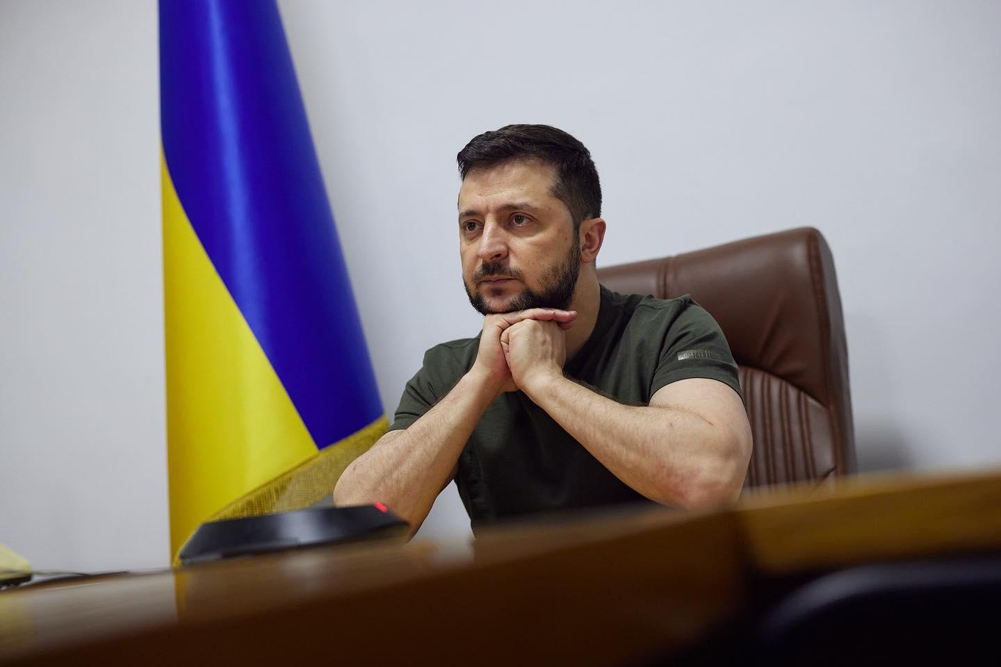 Volodymyr Zelensky sits at his desk with a Ukrainian flag behind him.