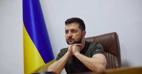 Volodymyr Zelensky sits at his desk with a Ukrainian flag behind him.