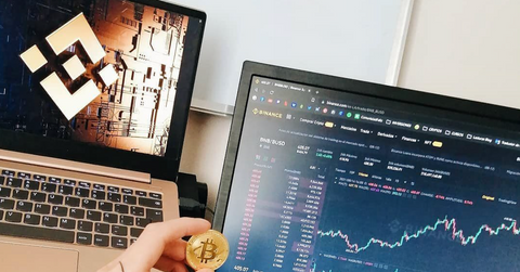 Someone holding a gold Bitcoin in front of a monitor displaying the Binance interface