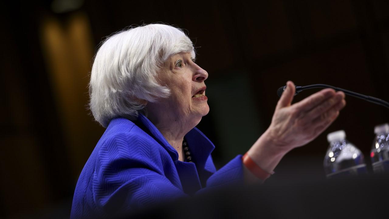 Janet Yellen in a blue shirt speaking at a podium.