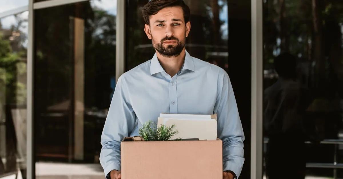 A man in a blue shirt leaves work with belongings after getting laid off.