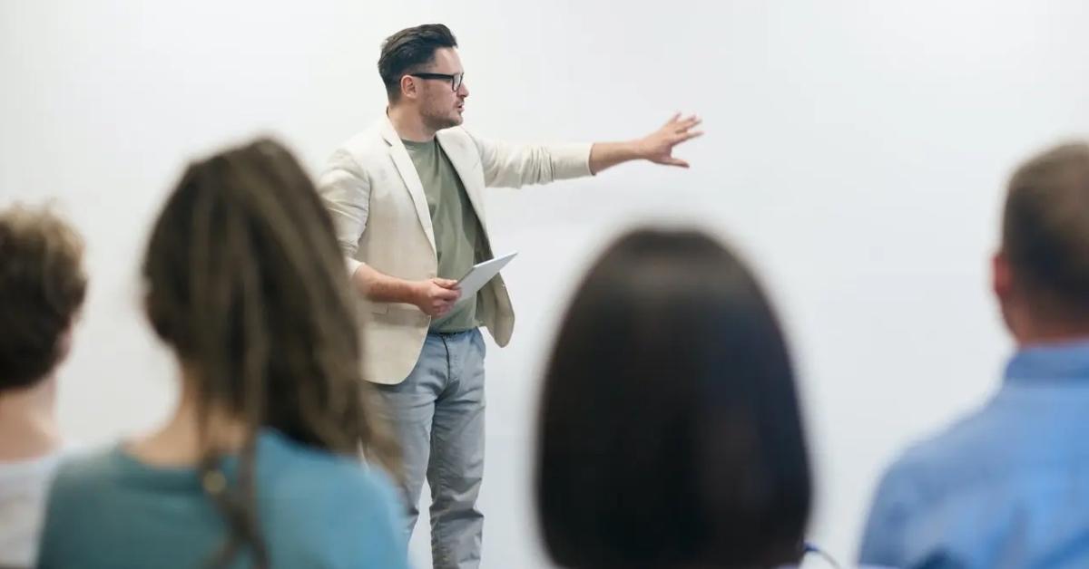 A male teacher in an ivory jacket lecturing a class.