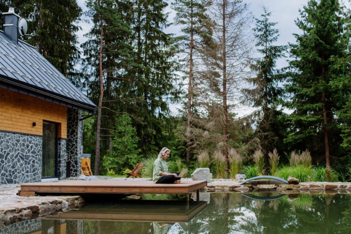 A woman sitting outside of a lake house with her laptop and headphones