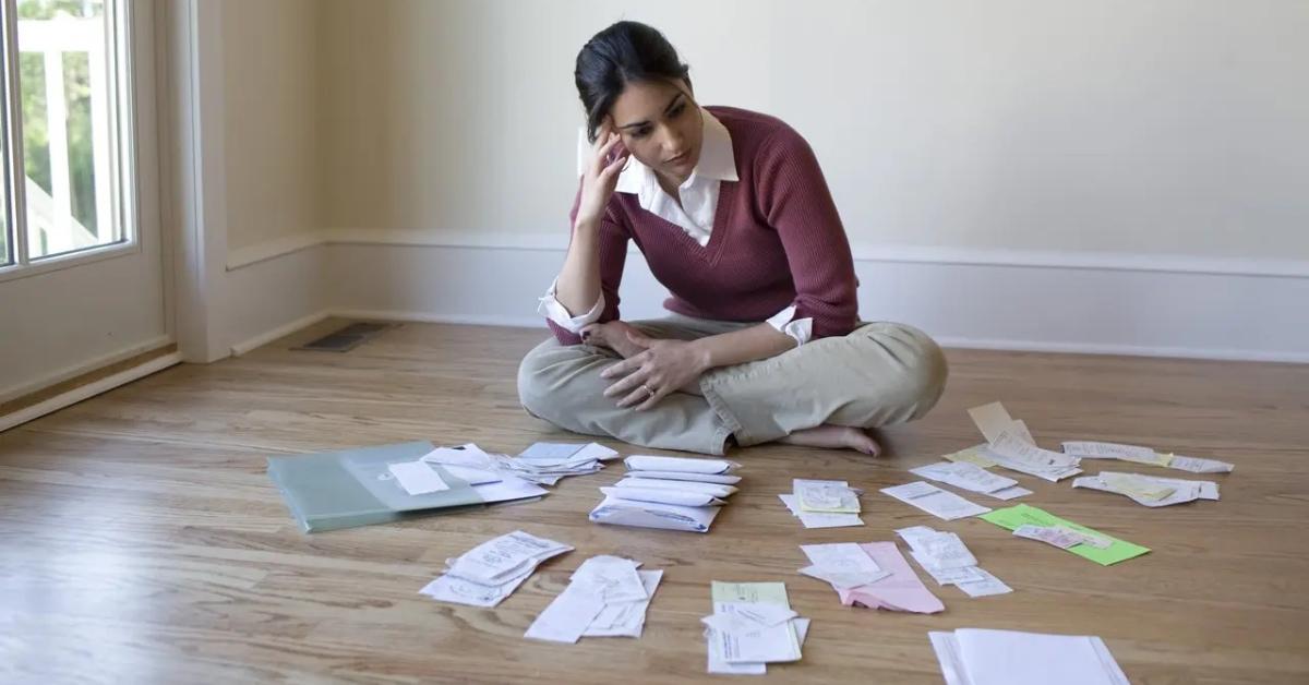 An overwhelmed woman in a maroon sweatshirt and khakis sitting on the floor surrounded by receipts.