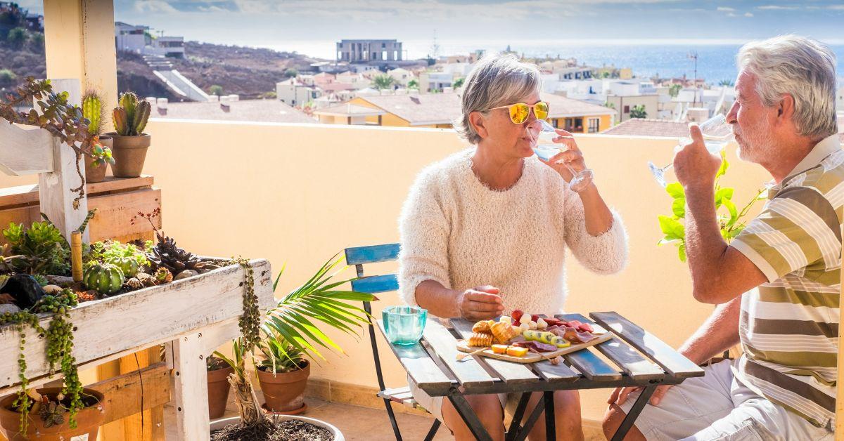 Two retirees sitting on a balcony with a cheese plate. 